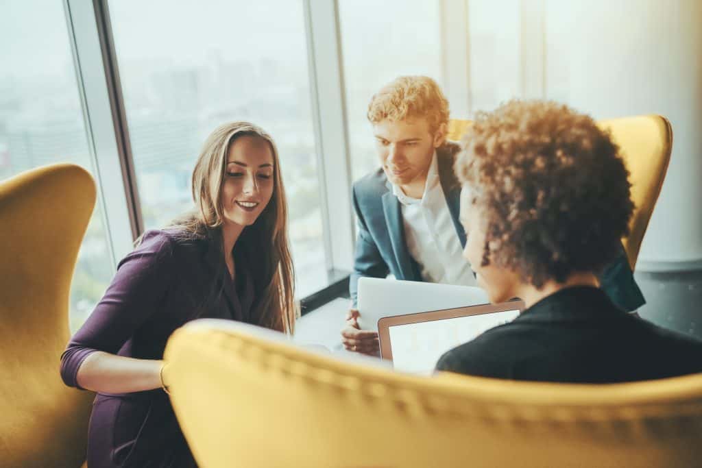 Attractive smiling businesswoman is showing screen of her gadget to her boss and female colleague during work meeting while sitting on yellow armchair in office interior next to window of skyscraper